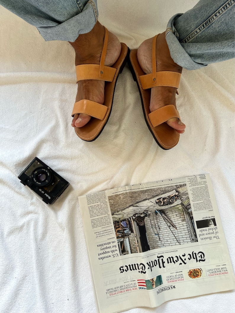May include: A pair of brown leather sandals with two straps across the foot. The sandals are worn by a person wearing blue jeans. The sandals are on a white surface.