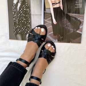 K&ouml;nnte beinhalten: Ein Paar schwarze Ledersandalen mit Kn&ouml;chelriemen. Die Sandalen haben ein geflochtenes Design und werden an den F&uuml;&szlig;en einer Person getragen.