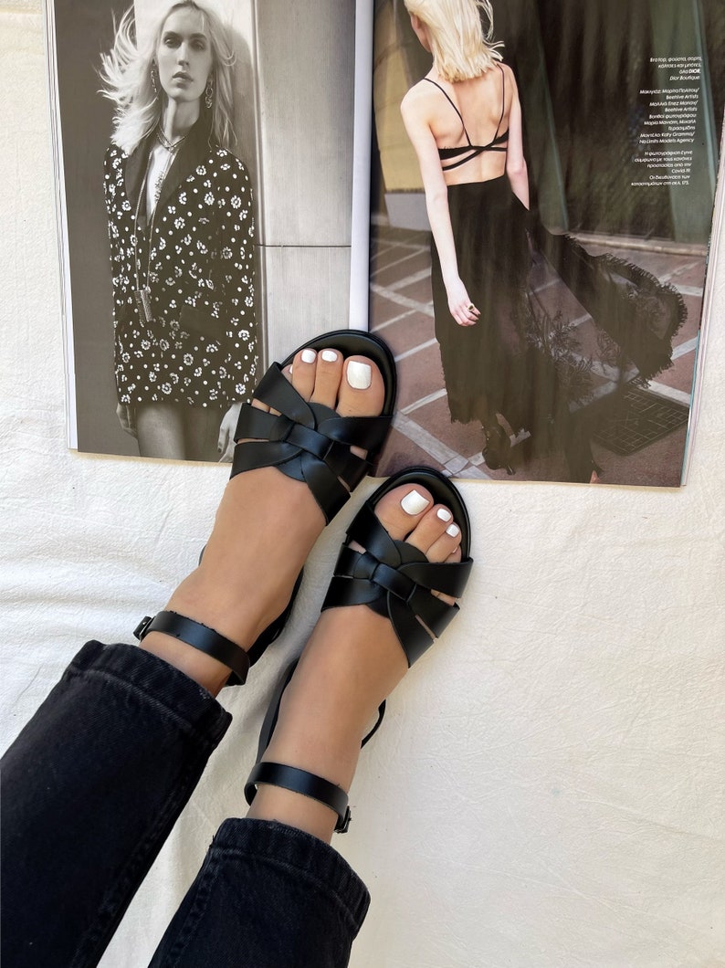 K&ouml;nnte beinhalten: Ein Paar schwarze Ledersandalen mit Kn&ouml;chelriemen. Die Sandalen haben ein geflochtenes Design und werden an den F&uuml;&szlig;en einer Person getragen.