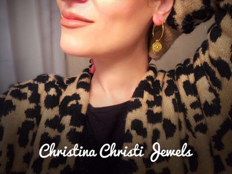 Puede incluir: Pendientes de aro dorados con un dije circular. Los pendientes se llevan con un su&eacute;ter con estampado de leopardo. El texto "Christina Christi Jewels" se muestra en la parte inferior de la imagen. Joyas.