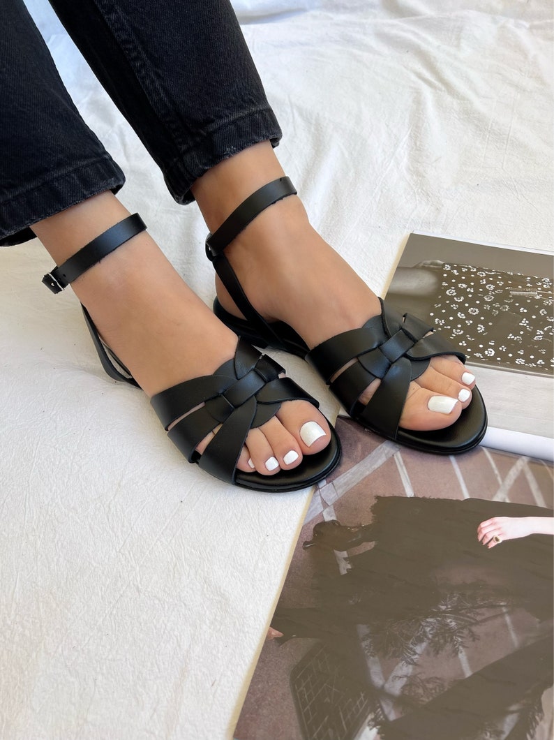 Puede incluir: Un par de sandalias de cuero negro con correas de tobillo y un dise&ntilde;o de cruz en el empeine. Las sandalias est&aacute;n puestas en los pies de una persona.