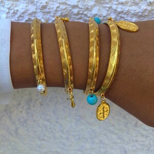Pode incluir: Quatro pulseiras de ouro com padr&otilde;es decorativos, cada uma com um amuleto diferente. Os amuletos incluem uma p&eacute;rola, uma conta turquesa e um pingente de cruz.