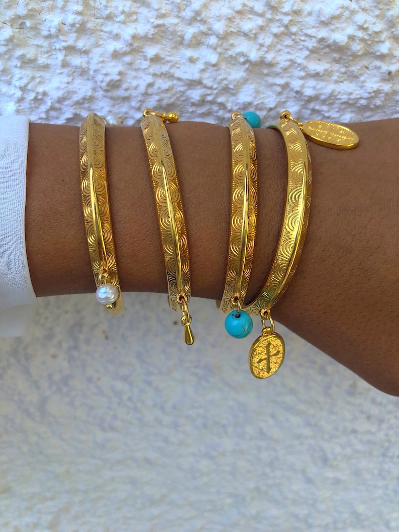 Pode incluir: Quatro pulseiras de ouro com padr&otilde;es decorativos, cada uma com um amuleto diferente. Os amuletos incluem uma p&eacute;rola, uma conta turquesa e um pingente de cruz.