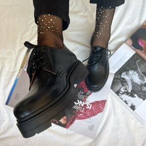 May include: Black leather lace-up platform shoes with a chunky sole. The shoes are worn with black sheer socks with silver glitter accents.