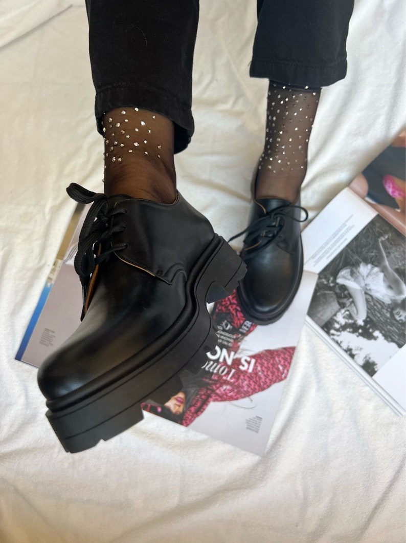 May include: Black leather lace-up platform shoes with a chunky sole. The shoes are worn with black sheer socks with silver glitter accents.