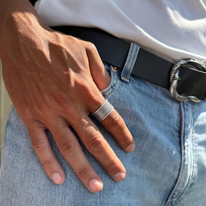 May include: A silver ring with multiple stacked bands is worn on a finger. The ring is on a hand, with a white t-shirt and blue jeans in the background. A black belt with a decorative buckle is also visible.