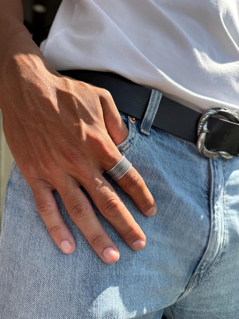May include: A silver ring with multiple stacked bands is worn on a finger. The ring is on a hand, with a white t-shirt and blue jeans in the background. A black belt with a decorative buckle is also visible.