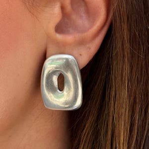 May include: A silver-coloured, rectangular earring with a rounded shape and a central oval opening. The earring is worn on an ear, with brown hair visible in the background. The earring has a smooth, reflective surface.