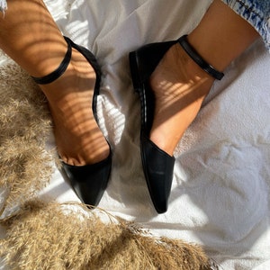 May include: A pair of black leather ankle strap flats with pointed toes. The shoes are on a white surface with a brown plant in the foreground.