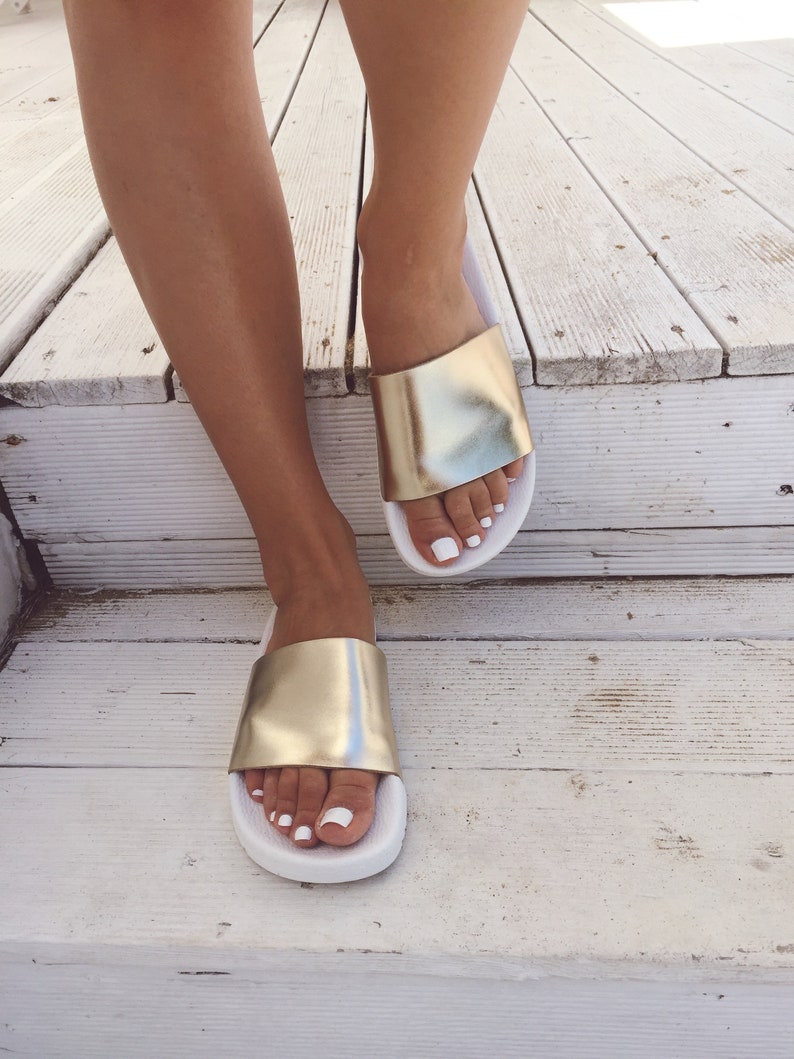 May include: A pair of white and gold slide sandals with a gold metallic strap. The sandals are on a wooden surface.