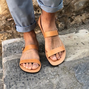 May include: Tan leather sandals with two straps and a buckle closure. The sandals are worn on a person's feet.