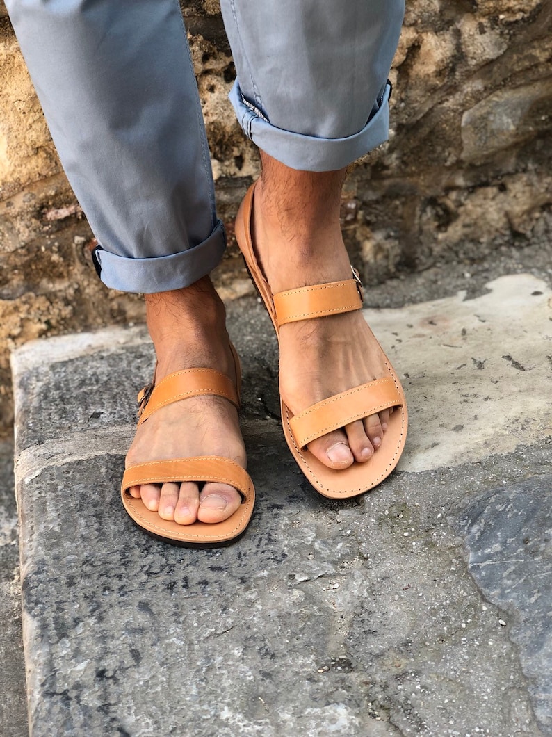 May include: Tan leather sandals with two straps and a buckle closure. The sandals are worn on a person's feet.
