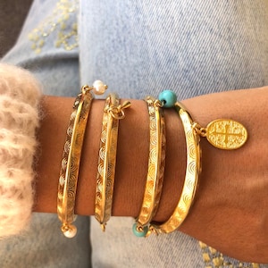 Pode incluir: Um conjunto de quatro pulseiras douradas com designs intrincados. As pulseiras est&atilde;o empilhadas no pulso de uma pessoa. Duas das pulseiras t&ecirc;m um pequeno charme de p&eacute;rola, e uma tem um charme turquesa. A quarta pulseira tem um charme de ouro com uma inscri&ccedil;&atilde;o.