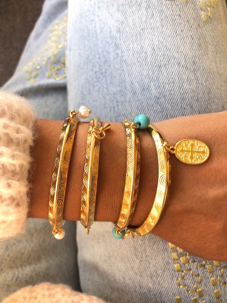 Pode incluir: Um conjunto de quatro pulseiras douradas com designs intrincados. As pulseiras est&atilde;o empilhadas no pulso de uma pessoa. Duas das pulseiras t&ecirc;m um pequeno charme de p&eacute;rola, e uma tem um charme turquesa. A quarta pulseira tem um charme de ouro com uma inscri&ccedil;&atilde;o.