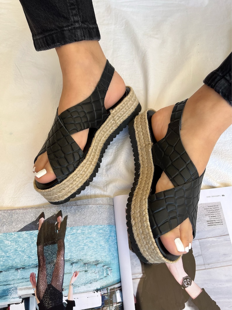 May include: Black platform sandals with a crisscross design and ankle strap. The sandals have a textured, faux-leather upper and a jute-wrapped platform sole. The sandals are shown on a white surface.