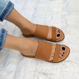 May include: A pair of brown leather sandals with two straps across the top of the foot. The sandals have a flat sole and are worn by a person with bare feet.
