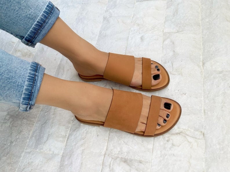 May include: A pair of brown leather sandals with two straps across the top of the foot. The sandals have a flat sole and are worn by a person with bare feet.