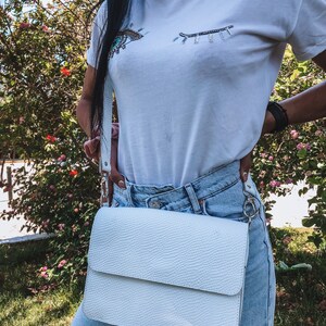 Puede incluir: Bolso bandolera de cuero blanco con superficie texturizada y cierre de solapa. El bolso se lleva sobre el hombro de una persona que lleva una camiseta blanca con un dise&ntilde;o de ojo de cuentas y unos vaqueros azules claros.