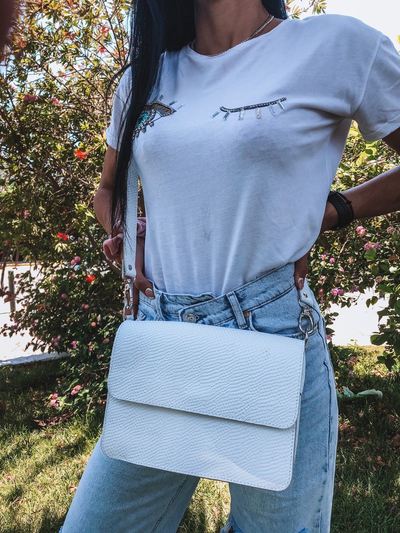 Puede incluir: Bolso bandolera de cuero blanco con superficie texturizada y cierre de solapa. El bolso se lleva sobre el hombro de una persona que lleva una camiseta blanca con un dise&ntilde;o de ojo de cuentas y unos vaqueros azules claros.
