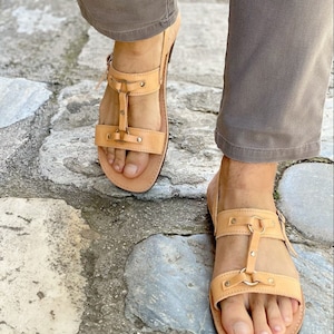 May include: Tan leather sandals with two straps and a buckle closure. The sandals have a  distressed look and are worn on a person's feet.