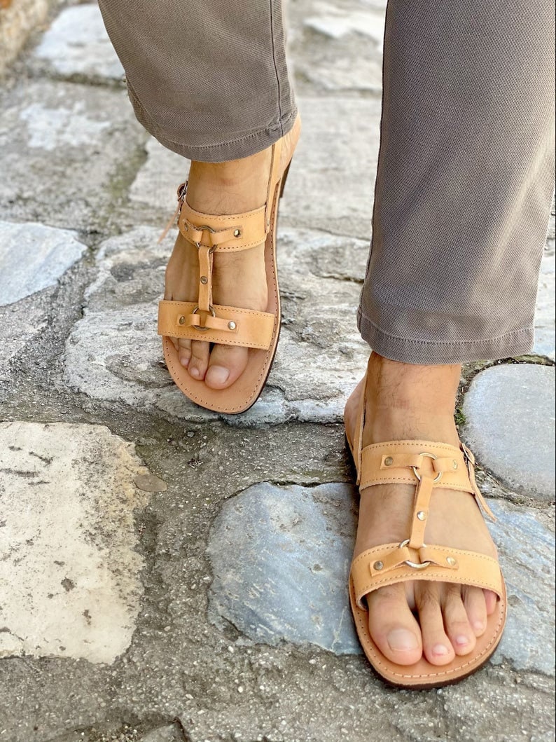 May include: Tan leather sandals with two straps and a buckle closure. The sandals have a  distressed look and are worn on a person's feet.