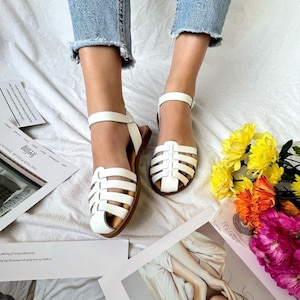 May include: White leather sandals with a woven strap design. The sandals have a buckle closure and a flat sole.