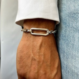 K&ouml;nnte beinhalten: Ein silbernes Kettenarmband mit einem gro&szlig;en, ovalen Glied. Das Armband besteht aus zwei verschiedenen Kettenarten, einer mit einem dickeren, massiveren Glied und einer mit einem d&uuml;nneren, filigraneren Glied.