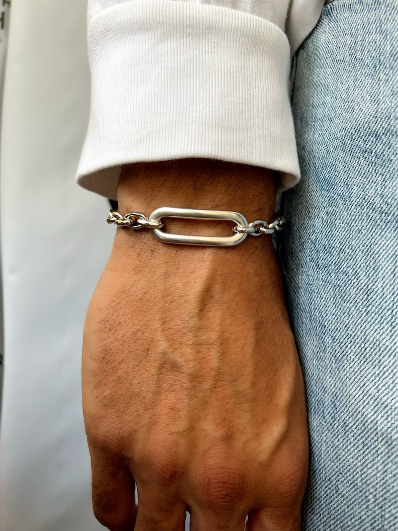 K&ouml;nnte beinhalten: Ein silbernes Kettenarmband mit einem gro&szlig;en, ovalen Glied. Das Armband besteht aus zwei verschiedenen Kettenarten, einer mit einem dickeren, massiveren Glied und einer mit einem d&uuml;nneren, filigraneren Glied.