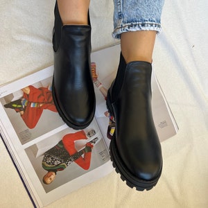 Pu&ograve; includere: Stivaletti in pelle nera con suola spessa. Gli stivaletti hanno pannelli laterali elasticizzati e sono indossati con jeans blu. Gli stivaletti sono appoggiati su una rivista.