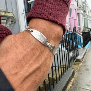 Puede incluir: Un brazalete de plata con un dije ovalado martillado y un diseño de cadena.