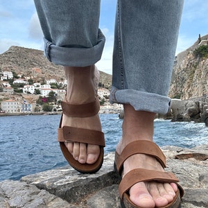 May include: Brown leather sandals with adjustable straps. The sandals have a cork footbed and are worn with blue jeans. The image is taken from a low angle, with a coastal town and sea in the background.