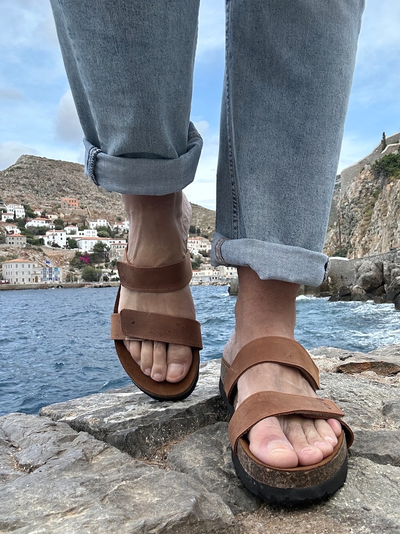 May include: Brown leather sandals with adjustable straps. The sandals have a cork footbed and are worn with blue jeans. The image is taken from a low angle, with a coastal town and sea in the background.