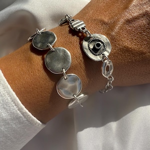 May include: Silver bracelet featuring a series of circular and oval links. The bracelet has a toggle clasp and a mix of smooth and textured surfaces. The design includes a central circular element with a dark center.