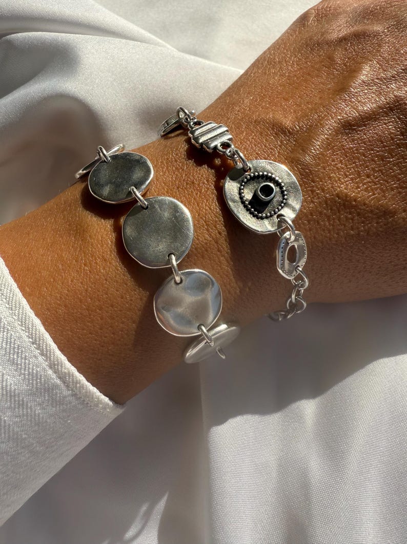 May include: Silver bracelet featuring a series of circular and oval links. The bracelet has a toggle clasp and a mix of smooth and textured surfaces. The design includes a central circular element with a dark center.