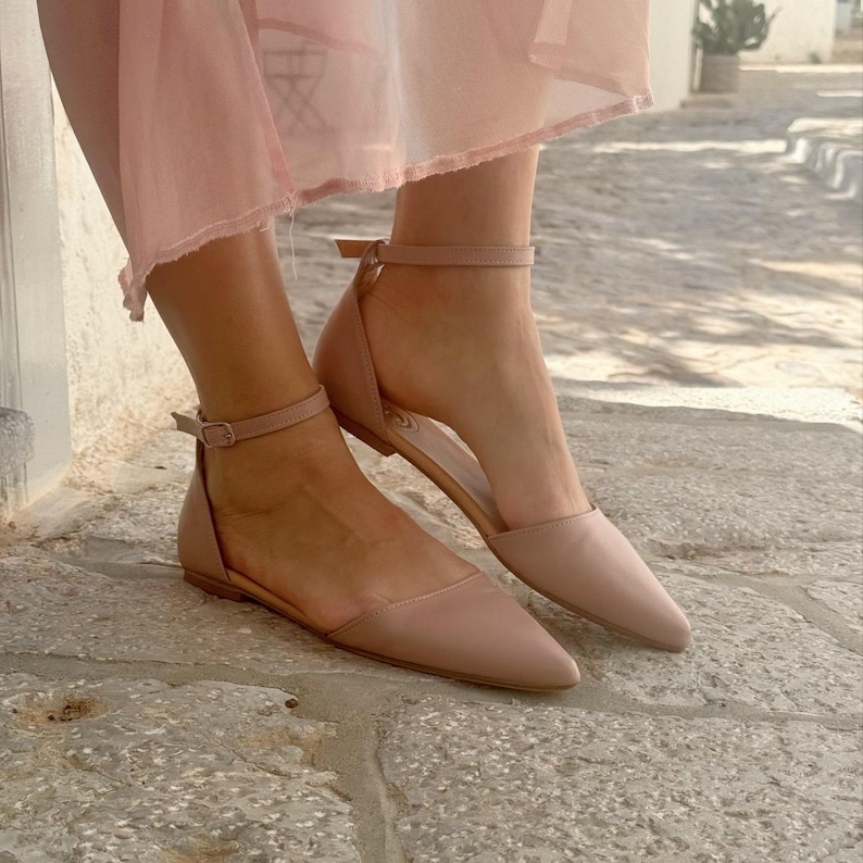 May include: A pair of blush-colored, pointed-toe flats with ankle straps. The shoes have a low heel and a simple, elegant design. The flats are made of a smooth material and are suitable for casual or dressy occasions.