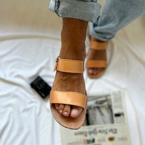 May include: Tan leather sandals with two straps and a buckle closure. The sandals have a simple design and are made of high-quality leather.