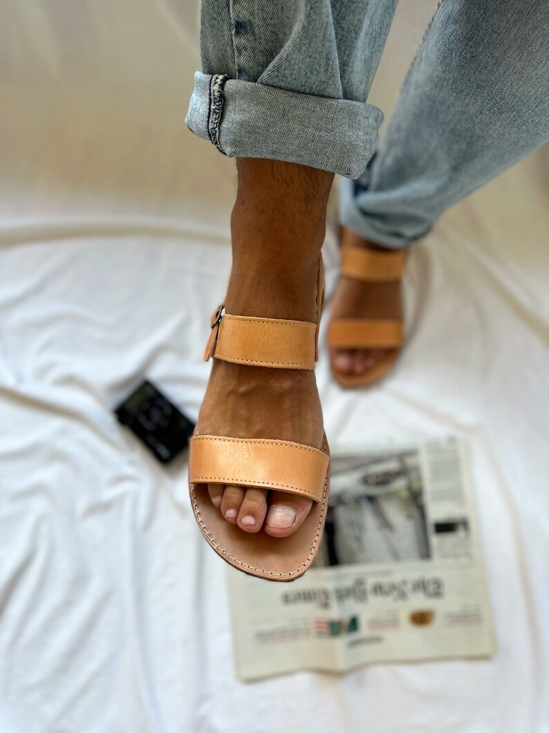 May include: Tan leather sandals with two straps and a buckle closure. The sandals have a simple design and are made of high-quality leather.