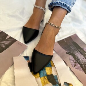 May include: Black pointed-toe flats with silver chain ankle straps. The shoes are paired with light blue denim jeans. The image is taken from a low angle, with art prints scattered around the feet.