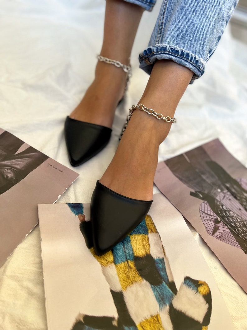May include: Black pointed-toe flats with silver chain ankle straps. The shoes are paired with light blue denim jeans. The image is taken from a low angle, with art prints scattered around the feet.