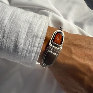 Könnte beinhalten: Ein silbernes Armband mit zwei rechteckigen, dunkelgrauen Paneelen, die durch eine Reihe kleiner, verbundener Stäbe verbunden sind. Das obere Paneel zeigt ein rot-oranges, strukturiertes Design in einem silbernen Rahmen.