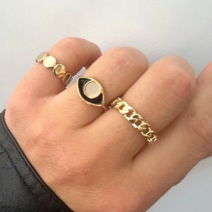 May include: Three gold rings on a hand. The ring on the index finger is a black and white eye design. The ring on the middle finger is a chain link design. The ring on the little finger is a row of small circles.