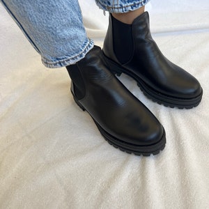 Pu&ograve; includere: Stivaletti Chelsea in pelle nera con suola spessa. Gli stivali hanno pannelli laterali elastici e sono indossati con jeans blu. Uno stile di calzatura classico.