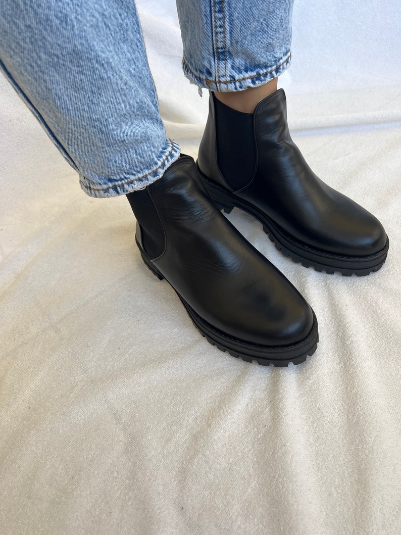 Pu&ograve; includere: Stivaletti Chelsea in pelle nera con suola spessa. Gli stivali hanno pannelli laterali elastici e sono indossati con jeans blu. Uno stile di calzatura classico.