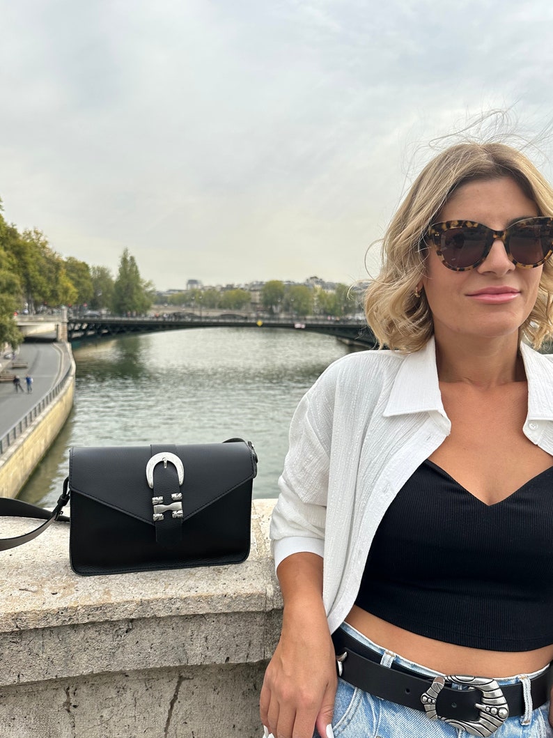 Puede incluir: Una mujer lleva una camisa blanca, un top negro y unos vaqueros azules con un cintur&oacute;n de hebilla plateada. Lleva un bolso bandolera de cuero negro con una hebilla de herradura plateada. El bolso cuelga de su hombro. Est&aacute; de pie en un puente con un r&iacute;o al fondo.