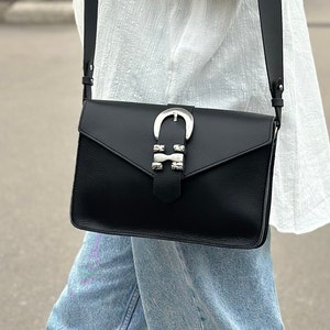 Puede incluir: Un bolso bandolera de cuero negro con una hebilla plateada y tachuelas. El bolso tiene cierre de solapa y una correa larga ajustable.