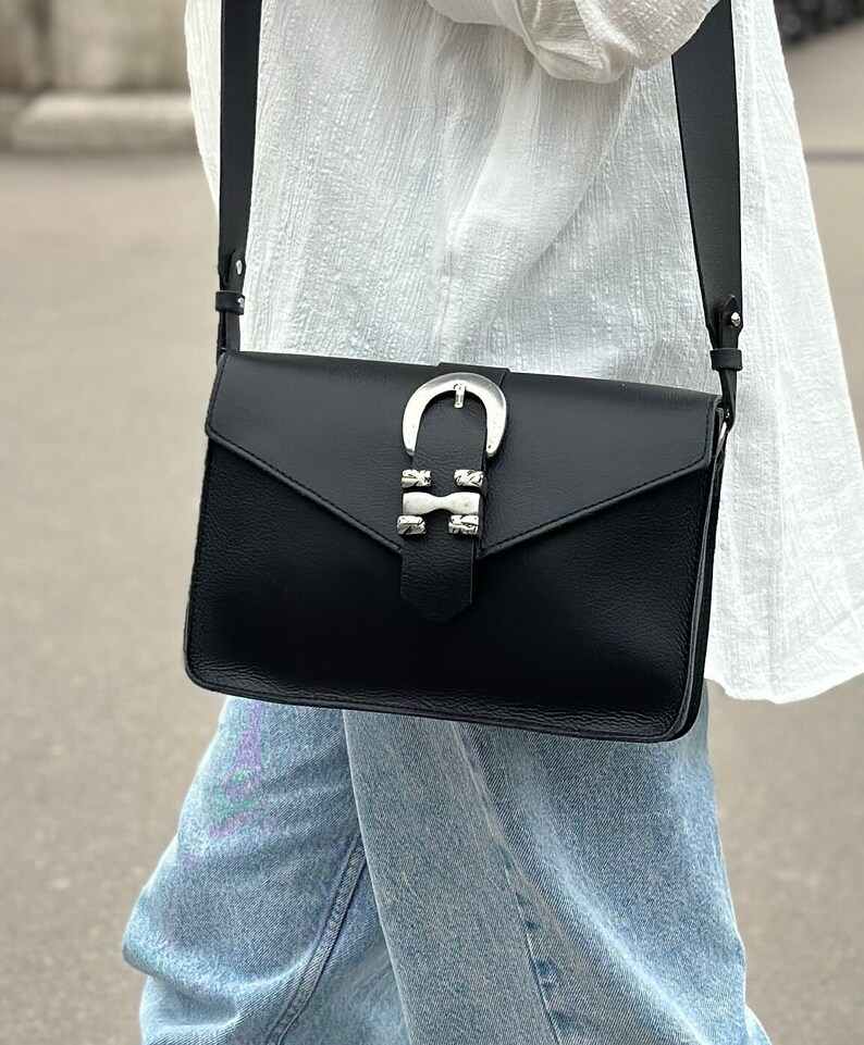 Puede incluir: Un bolso bandolera de cuero negro con una hebilla plateada y tachuelas. El bolso tiene cierre de solapa y una correa larga ajustable.