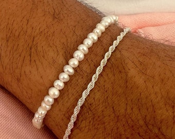 May include: A white pearl bracelet and a silver rope chain bracelet on a person's wrist.