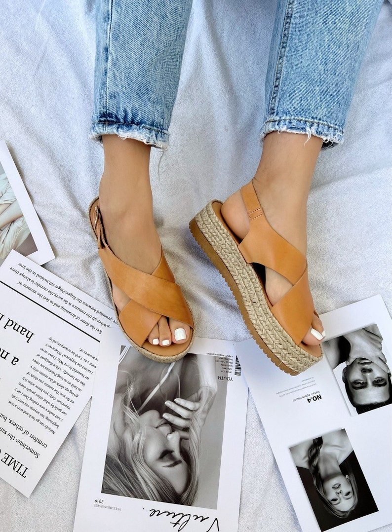 May include: Tan leather sandals with crisscross straps and a woven platform sole. The sandals are worn with blue jeans. Magazines with black and white photos are scattered around the feet.