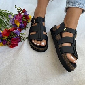 May include: Black leather sandals with a cork platform sole and multiple straps. The sandals have a buckle closure and gold accents.