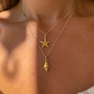 Op de afbeelding: Een gouden ketting met twee hangers: een zeester en een schelp. De ketting is gelaagd en de hangers zijn delicaat.